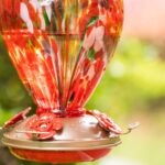 A vibrant hand-blown glass hummingbird feeder with a floral balloon design, hanging outdoors in a garden.