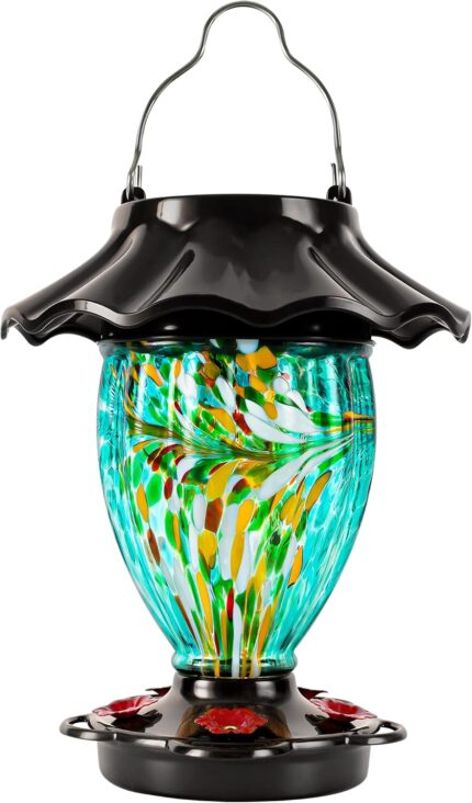 A teal hand-blown glass LUJII solar-powered hummingbird feeder hanging outdoors, glowing with color-changing light at dusk.
