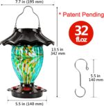 A teal hand-blown glass LUJII solar-powered hummingbird feeder hanging outdoors, glowing with color-changing light at dusk.