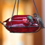 A vibrant red, hand-blown glass hummingbird feeder with a decorative hanging basket and three feeding ports.